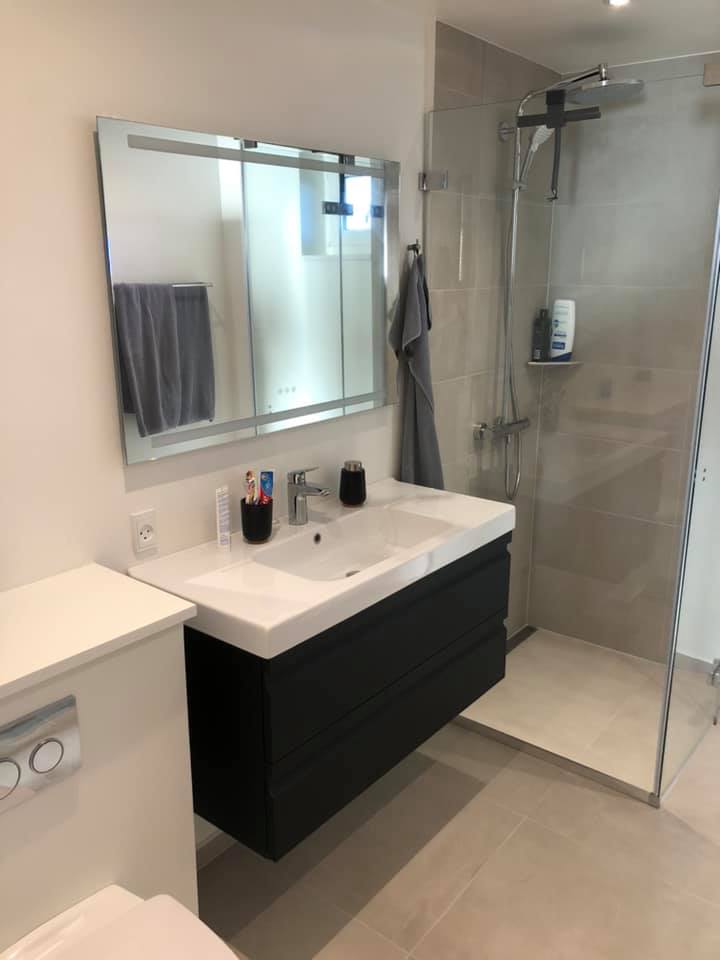 Lyst badeværelse med væghængt vaskeskab, bruseniche og lyse gulv- og vægfliser som eksempel på professionel opsætning af klinker.