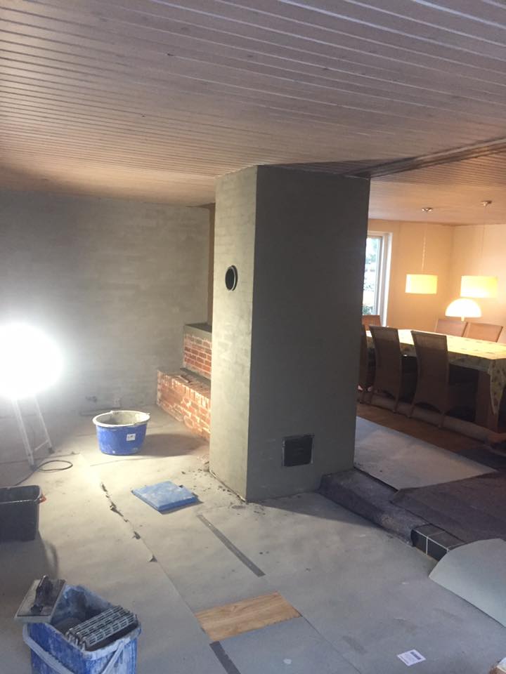 Stue under renovering med pudset skorsten i midten af rummet og byggematerialer på gulvet.