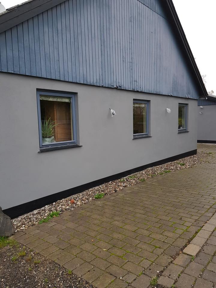 Hus med ny, jævn grå facade og blå vinduer langs brolagt indkørsel – resultat efter facaderenovering.