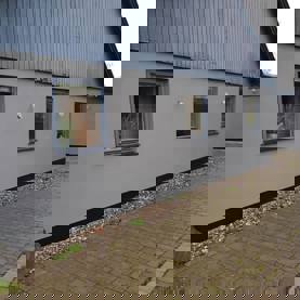 Hus med ny, jævn grå facade og blå vinduer langs brolagt indkørsel – resultat efter facaderenovering.