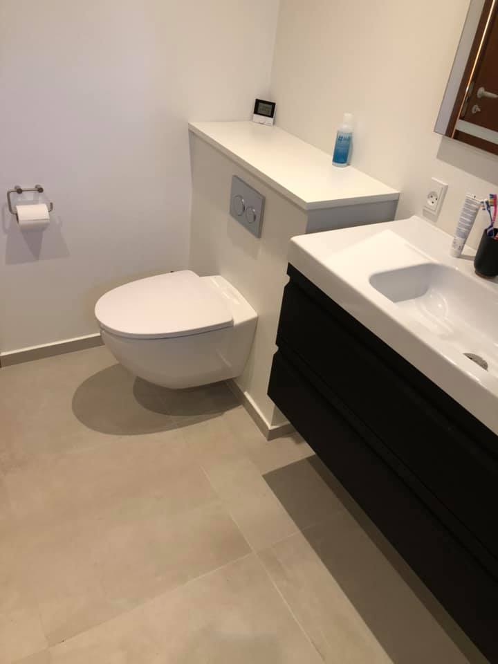 Lyst badeværelse med væghængt toilet, sort vaskeskab og lyse gulvklinker lagt i store rektangler.