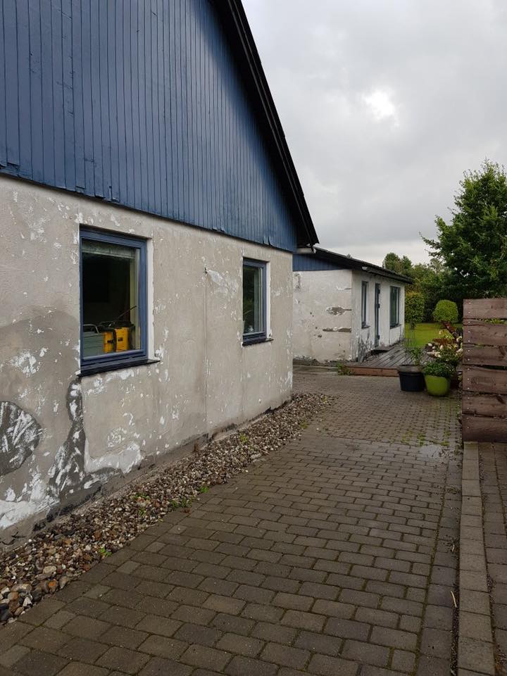 Hus med afrenset og ujævn grå facade langs brolagt indkørsel som er midt i facaderenovering.